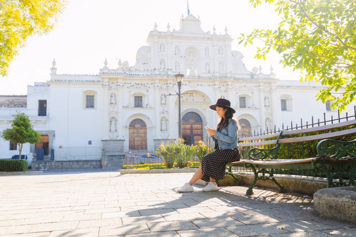 Young Hispanic woman sitting watching her phone in front of the San Jose cathedral in Antigua Guatemala-tourist traveling in colonial city-woman in the central park of Antigua Guatemala