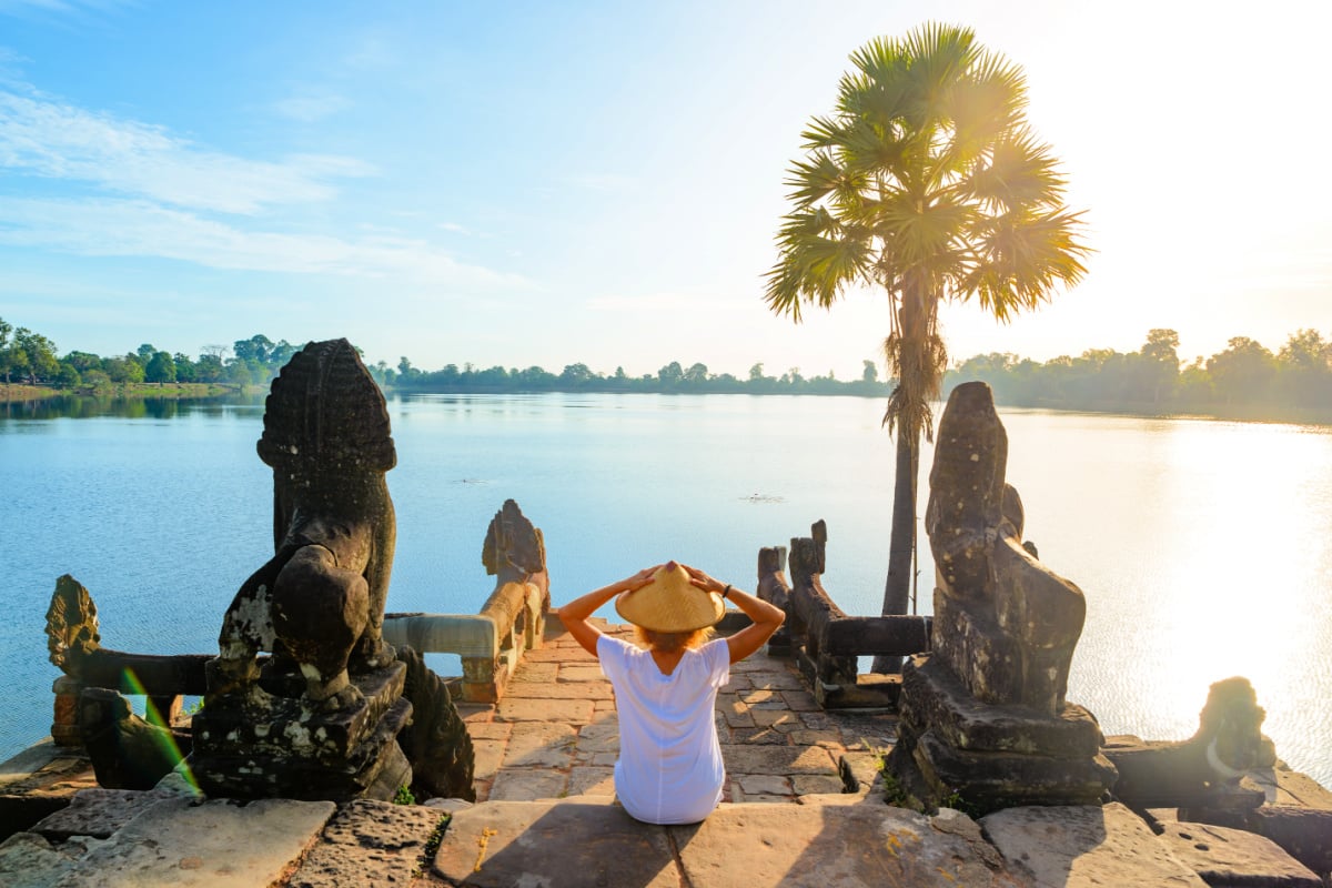 One tourist relaxing in Angkor ruins at sunrise, Srah Srang temple water pond amid jungle, travel destination Cambodia. Woman with traditional hat, rear view.